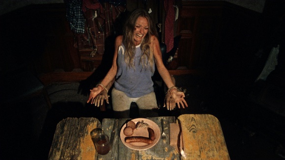 a screaming, injured woman is bound to the "arms" of a chair that appear to be actual human arms, at an old table in a ramshackle house