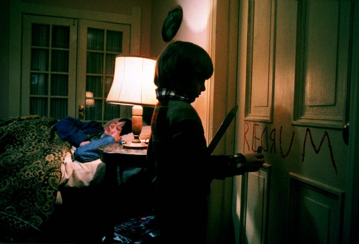 Danny Torrence writes "REDRUM" in red crayon on a door while holding a knife as his mother sleeps in bed in The Shining