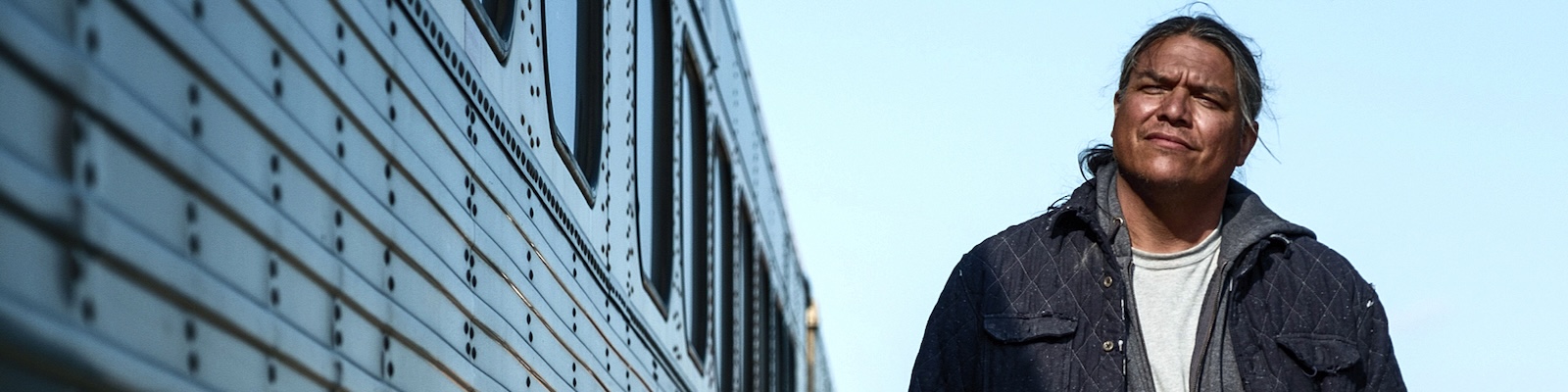 a man with long black hair smiles as he stands next to a train