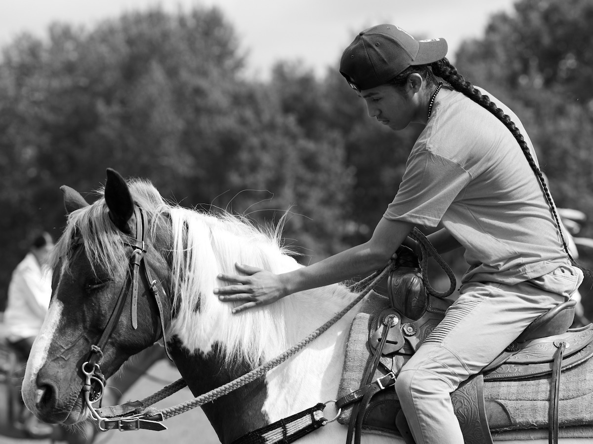 A young man on horseback with a long braid wears a baseball cap and tshirt. He is patting the horse's mane.