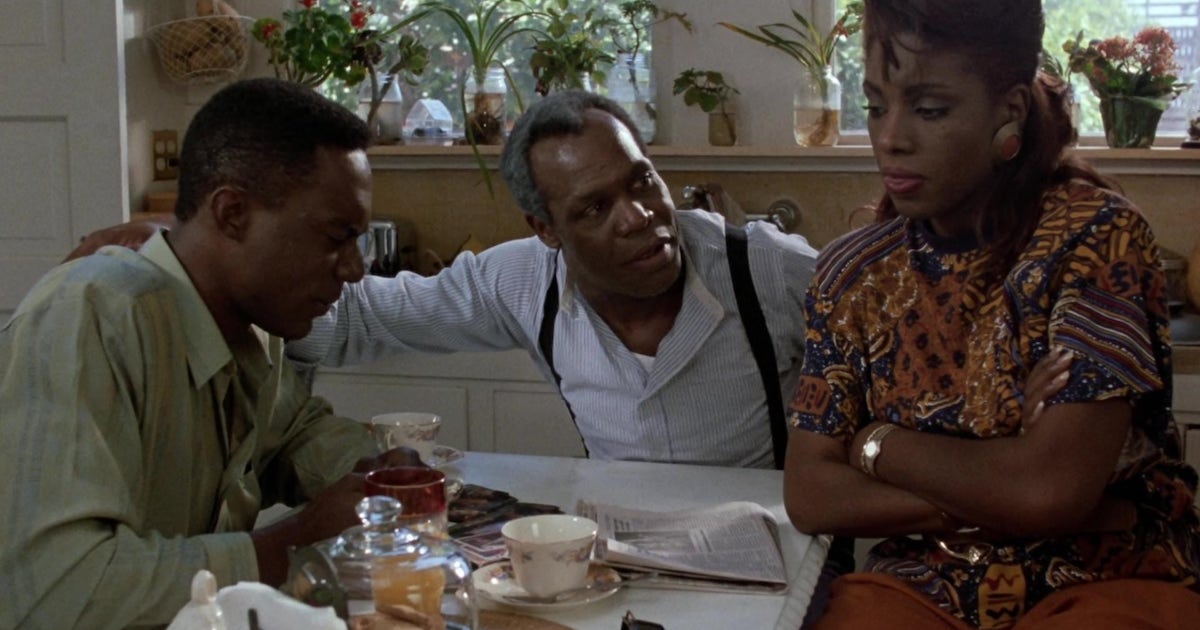 An older black man (Danny Glover) sits at the head of a small kitchen table covered with a newspaper and coffee cups. His arms are around a black woman and a young black man who sit on either side of him. Image from the film To Sleep With Anger.