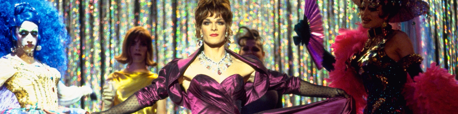 Patrick Swayze, a white man, appears in drag in the film To Wong Foo. He wears a brunette wig in an up-do, and a purple satin strapless gown with a matching shrug jacket. His arms are out stretched holding the skirt of the gown. He has long black lace gloves, and a chunky jewel necklace and earing. He is on a stage surrounded by out of focus drag queens.