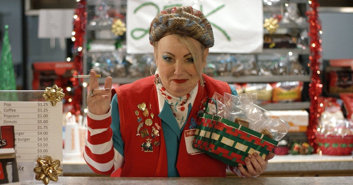 Jann Arden stands behind a concessions counter wearing a red vest covered in pins and a name tag, with a cigarette in one hand and a Christmas basket in the other. She has curlers in her hair and is wearing a lot of blue eye shadow and bright red lipstick.