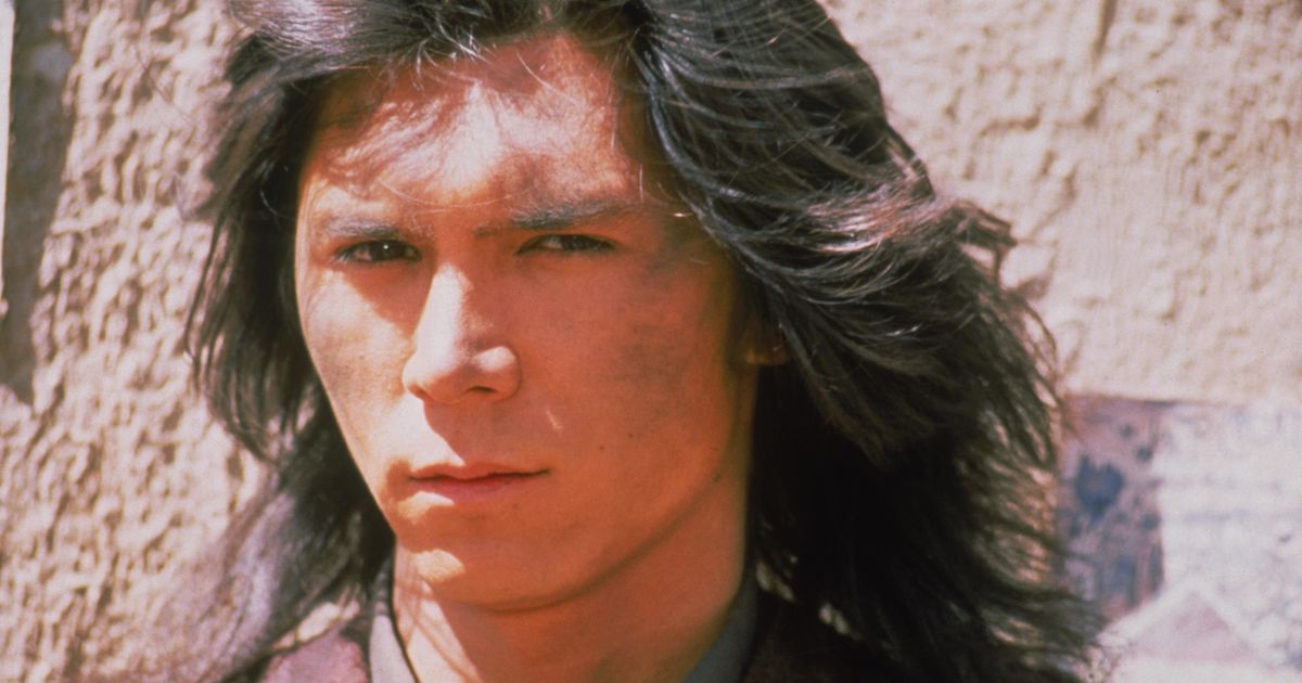 Close up of the face of Lou Diamond Phillips A mid-twenties mixed-race man with long black hair. His expression is stern and cool. There is a patch of dirt on his face. Promotional image from the film Young Guns